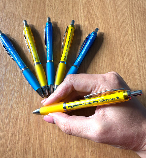 Hand holding a yellow pen with 'Together we make the difference' text, surrounded by similar pens on a wooden surface.